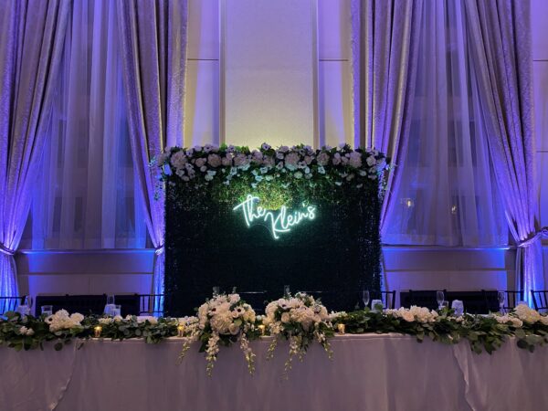 Wedding head table at The Inn at St. John’s Grand Ballroom with blue uplighting, a custom hedge wall, and a neon last name sign behind the newlyweds.